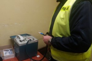 Na zdjęciu widać policjanta, który stoi obok urządzenia służącego do badania stanu trzeźwości. Policjant w ręku trzyma rurkę zakończoną ustnikiem.