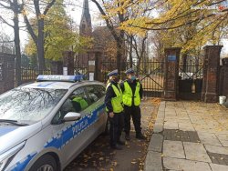 Dwóch policjantów stojących przy radiowozie obserwuje cmentarz.