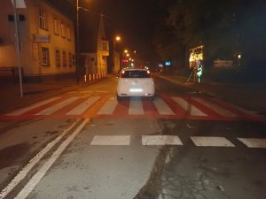 Na zdjęciu widać samochód osobowy na drodze, który zatrzymał się w połowie przejścia dla pieszych. W tle zdjęcia widać policjanta wykonującego pomiary