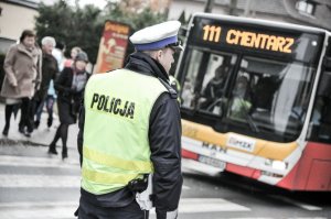 Na zdjęciu widać policjanta z drogówki znajdującego się przy przejściu dla pieszych. Po przejściu przechodzą ludzie, którzy wysiedli z autobusu, na którym wyświetla się napis &quot;cmentarz&quot;.