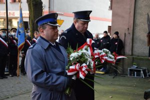 Komendant Powiatowy Policji w Tarnowskich Górach młodszy inspektor Sławomir Bylicki oraz przedstawiciel Aresztu Śledczego stoją z wiązankami kwiatów przed  tablicami pamięci.