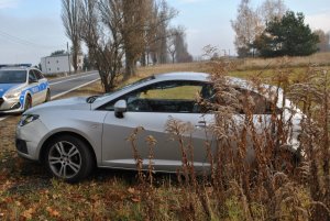 Na zdjęciu widać samochód osobowy w przydrożnym rowie. W tle zdjęcia widać policyjny radiowóz