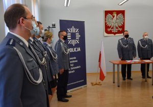 Przy oknie stoją policjanci w umundurowaniu galowym. W tle zdjęcia widać szefa i wiceszefa tarnogórskich policjantów.