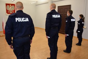 Czworo policjantów stojących obok siebie na baczność. Przed nimi znajduje się Godło Polski i Biało-Czerwona Flaga.