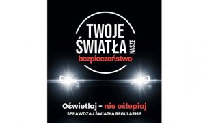 Na czarnym tle świecące przednie reflektory pojazdu, nad nimi napis „Twoje światła nasze bezpieczeństwo”. Poniżej napis „Oświetlaj – nie oślepiaj, sprawdzaj światła regularnie.