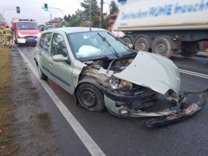 Na zdjęciu widać drogę, na której znajduje się rozbity samochód marki Renault. W tle zdjęcia widać wóz strażacki i strażaków oraz przejeżdżającą ciężarówkę
