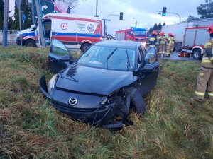Na zdjęciu widać rozbity samochód osobowy marki Mazda, który znajduje się w przydrożnym rowie. Obok auta stoi strażak. W tle widać ambulans.