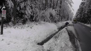 Na środku drogi biegnącej przez las znajduje się laweta. W przydrożnym rowie leży pojazd. Okolica jest cała zaśnieżona.