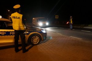Na zdjęciu widać dwóch policjantów znajdujących się w rejonie przejścia dla pieszych. Jeden stoi na poboczu drogi obok radiowozu, drugi zatrzymuje do kontroli drogowej kierowcę busa.