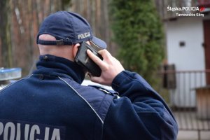 Na zdjęciu widać policjanta stojącego przed ogrodzeniem posesji. Policjant rozmawia przez telefon komórkowy z osobą objętą kwarantanną