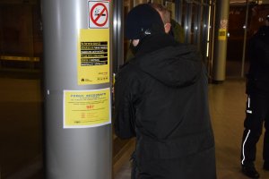 Pracownik socjalny na słupie dworca autobusowego klei plakat z informacjami dotyczącymi pomocy dla osób bezdomnych.