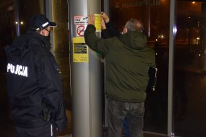 Pracownik socjalny na słupie dworca autobusowego klei plakat z informacjami dotyczącymi pomocy dla osób bezdomnych. Obok niego znajduje się policjant.