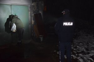 Policjant i pracownik socjalny stoją przy drzwiach budynku, w którym przebywają osoby bezdomne. Pracownik trzyma w ręku koc i kołdrę.