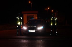Na zdjęciu widać dwóch policjantów stojących obok radiowozu. Policjanci mają na sobie elementy odblaskowe, które odbijają się w świetle.