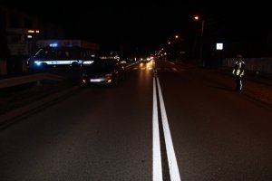 Na zdjęciu widać drogę z oznakowanym przejściem dla pieszych. Po jednej stronie drogi stoi policjant, a po drugiej znajduje się policyjny radiowóz. Przed przejściem znajduje się samochód biorący udział w zdarzeniu.