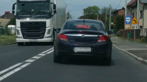 Na zdjęciu widać jezdnię rozdzieloną liniami. Po jezdni jedzie nieoznakowany policyjny radiowóz. W radiowozie, w tylnej szybie wyświetla się czerwony napis kontrola. Z przeciwnego kierunku jedzie samochód ciężarowy, który mija się z radiowozem.
