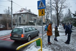 Na zdjęciu widać przejście dla pieszych. przez które przejeżdża samochód. Obok przejścia znajduje się policjant, który rozmawia z przechodzącymi przez przejście osobami.