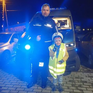 Na zdjęciu widać policjanta, który stoi obok chłopca ubranego w kamizelkę odblaskową. Z tyłu znajduje się policyjny radiowóz.