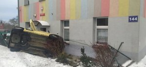 Na zdjęciu widać leżący na boku samochód osobowy. Auto opiera się o elewację budynku.  Widać również zniszczone ogrodzenie znajdujące się przed budynkiem.