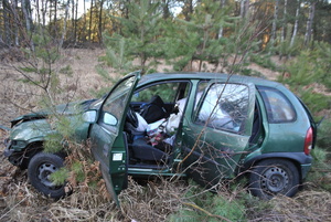 Na zdjęciu widać zniszczone auto znajdujące się w przydrożnym rowie. Samochód ma uszkodzoną karoserię, otwarte drzwi od strony kierowcy i pasażera.