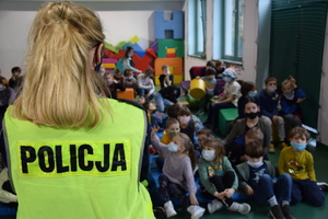 na zdjęciu widoczna policjantka stojąca tyłem, w niebieskim mundurze i kamizelce odblaskowej z napisem policja, w tle dzieci siedzące na materacach w sali