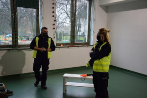na zdjęciu policjant i policjantka w niebieskich mundurach i kamizelkach odblaskowych w sali szkolnej, pomiędzy nimi biały stolik a na nim policyjna latarka z pomarańczowym elementem i tarcza do zatrzymywania pojazdów, w tle dwa okna, widok na podwórko
