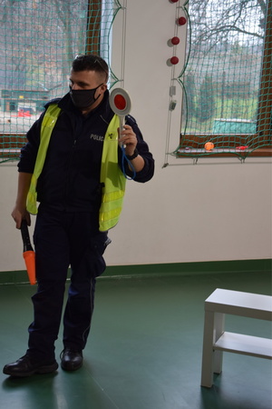 na zdjęciu policjant w niebieskim mundurze i kamizelce odblaskowej, w prawej ręce trzyma policyjną latarkę, a w lewej tarczę do zatrzymywania pojazdów, w tle okno z widokiem na podwórko