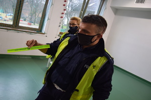 na zdjęciu policjant w niebieskim mundurze i kamizelce odblaskowej trzymający w prawej ręce opaskę odblaskową koloru zielonego z napisem powiat tarnogórski, herbem powiatu oraz stroną internetową, w tle policjantka, okna z widokiem na podwórko