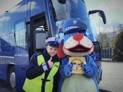 na zdjęciu widocznych policjant w niebieskim mundurze i odblaskowej kamizelce, w białej czapce, obok maskotka policji Sznupek, w tle niebieski autobus