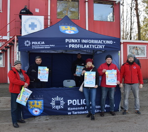 na zdjęciu namiot koloru niebieskiego z napisem komenda powiatowa policji w tarnowskich górach oraz herbem i napisem starostwo powiatowe, w namiocie dwóch policjantów ubranych z niebieskie mundury, trzymający w ręku kolorowy plakat, przed namiotem trzech mężczyzn w czerwonych polarach oraz kobieta w niebieskich spodniach, czarnej kurtce, bordowej czapce, kobieta i dwóch mężczyzn trzyma w ręku kolorowy plakat, w dalszym planie czerwony, piętrowy budynek, z białą tablicą, na której widnieje niebieski krzyż i napis wodne ochotnicze pogotowie ratunkowe, do budynku na drugim planie wchodzi policjant w niebieskim mundurze z napisem policja na plecach