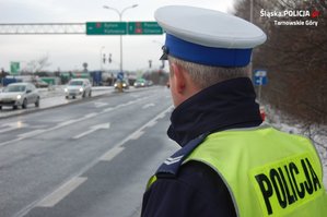 na zdjęciu policjant w niebieskim mundurze, kamizelce odblaskowej, białej czapce, obserwuje widoczną w tle jezdnie