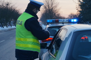 na zdjęciu policjant w niebieskim mundurze, odblaskowej kamizelce i białej czapce w trakcie kontroli pojazdu, w tle radiowóz