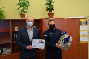 na zdjęciu z lewej strony kierownik rewiru dzielnicowych w jasnym garniturze trzymający w ręku zdjęcie z podziękowaniami, obok w niebieskim mundurze naczelnik wydziału prewencji z koszem słodkości