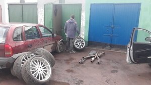 z lewej strony samochód osobowy koloru ciemnego, oparte o niego koła, w centralnej części mężczyzna w ciemnym ubiorze stojący przed budynkiem, jedne z drzwi budynku otwarte, z prawej strony drzwi pojazdu osobowego
