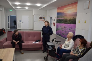 na zdjęciu widoczna policjantka w niebieskim mundurze stojąca przy sofie, na sofie po lewej i prawej stronie seniorzy słuchające wykładu policjantki, w tle fototapeta z kwiatami, drzwi pomieszczenia