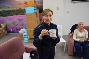 policjantka w niebieskim mundurze trzymająca w rękach kubek z napisem nie akceptujesz - reagujesz!, obok na sofie siedzącą kobieta w jasnym ubiorze, w tle kartony