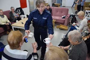 policjantka w niebieskim mundurze wręczająca kubki seniorom, z napisem nie reagujesz - akceptujesz. w tle seniorzy siedzący na sofie
