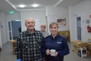 policjantka w niebieskim mundurze, obok mężczyzna w ciemnej koszuli w kratkę, policjantka trzyma kubek z napisem nie reagujesz - akceptujesz. w tle kartony i starszy mężczyzna