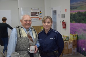 na zdjęciu policjantka w niebieskim mundurze, obok niej mężczyzna w niebieskiej koszuli, kamizelce i krawacie, trzymający w ręku kubek z napisem nie reagujesz - akceptujesz, w tle kartony i idąca kobieta
