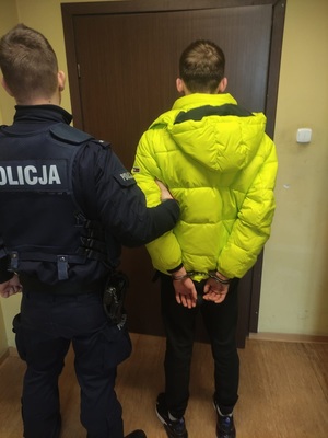 na zdjęciu policjant w niebieskim mundurze trzyma za rękę zatrzymanego, w żółtej kurtce, skutego kajdankami, w tle brązowe drzwi