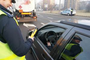 policjant w niebieskim mundurze, odblaskowej kamizelce i białej czapce trzyma w prawej ręce urządzenia do badania trzeźwości. stoi przy pojeździe koloru czarnego, kierowca ma otwartą szybę. w tle jezdnia, biała cysterna i radiowóz policyjny