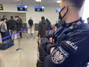 policjant z naszywką na lewym ramieniu z napisem &quot;komenda powiatowa policji&quot; obserwuje osoby w kolejce do odprawy, w terminalu lotniska