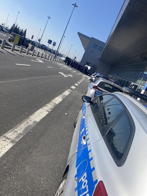 lewy bok policyjnego radiowozu, przed nim kolejne, w tle policjanci na zbiórce