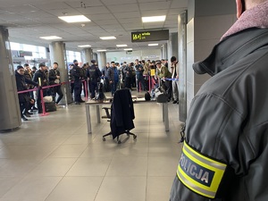 lewe ramię funkcjonariusza, który w ubiorze cywilnym trzyma na ramieniu opaskę odblaskową z napisem policja, w tle osoby w terminalu lotniska, obok policjanci w mundurach