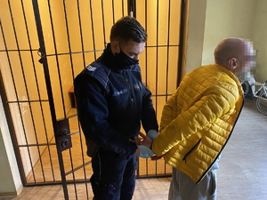 policjant w niebieskim mundurze zakłada kajdanki na ręce trzymane z tyłu zatrzymanemu, który ubrany jest w żółtą kurtkę i szare spodnie