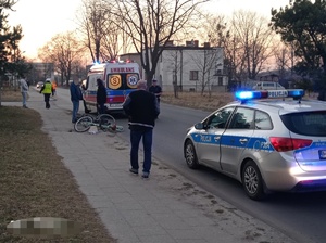na zdjęciu, po prawej stronie radiowóz zaparkowany na jezdni przy chodniku. Obok chodnikiem porusza się mężczyzna w ciemnym ubiorze, przed nim leżący rower koloru jasne, częściowo na jezdni. Następnie karetka pogotowia ratunkowego. Obok karetki z prawej strony policjant w mundurze i białej czapce, z prawej inne osoby. W tle zabudowania.