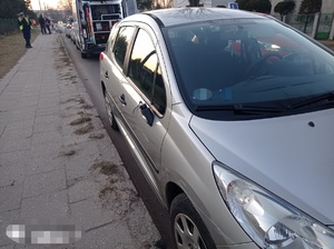 na pierwszym planie widzimy lewy bok pojzdu, w którym brakuje lusterka. W tle na chodniku policjant rozmawiający przez telefoh, obok radiowóz z otwartmi tylnymi drzwiami. w tle osoby i zabudowania. Leżący na chodzniku i jezdni rower.