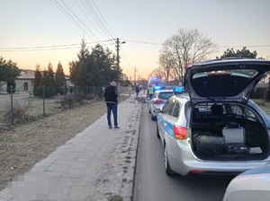 policyjne radiowozy zaparkowane jeden za drugim. Pierwszy ma otwarty bagażnik. Na chodniku stoi mężczyzna. W tle karetka pogotowia, osoby i zabudowania.