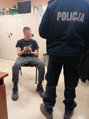 na zdjęciu policjant stoi obok siedzącego mężczyzny na krześle skutego w kajdanki, za nim białe szafy