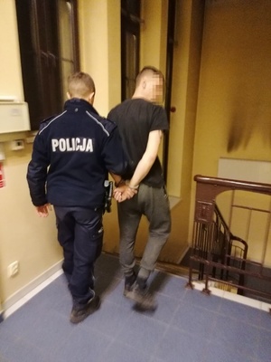policjant prowadzący mężczyznę skutego w kajdanki na ręce z tyłu,